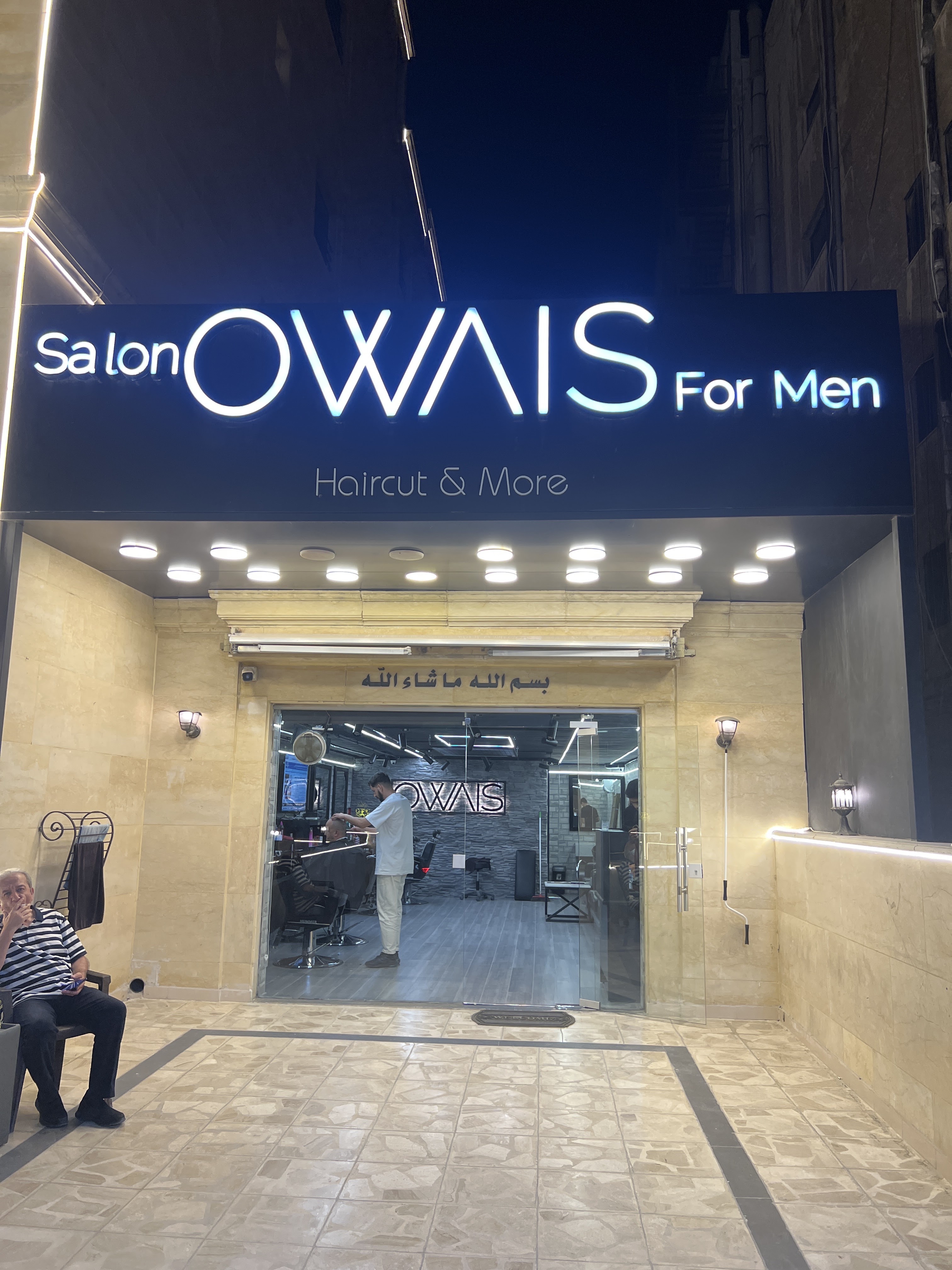 Salon Owais For Men storefront lit up at night with a barber working inside